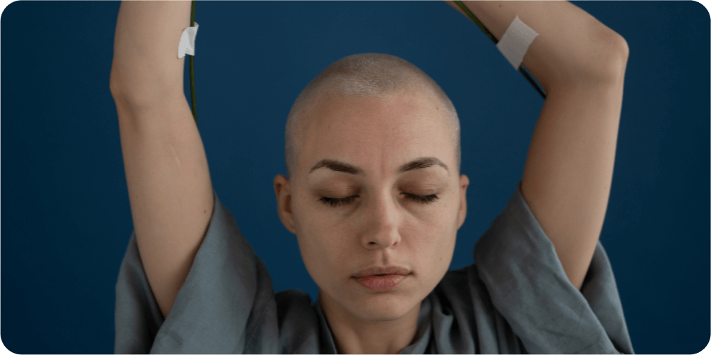 Спокойная лысая женщина с закрытыми глазами поднимает руки на синем фоне. На ней серый топ; на руках видны медицинские пластыри и трубки.