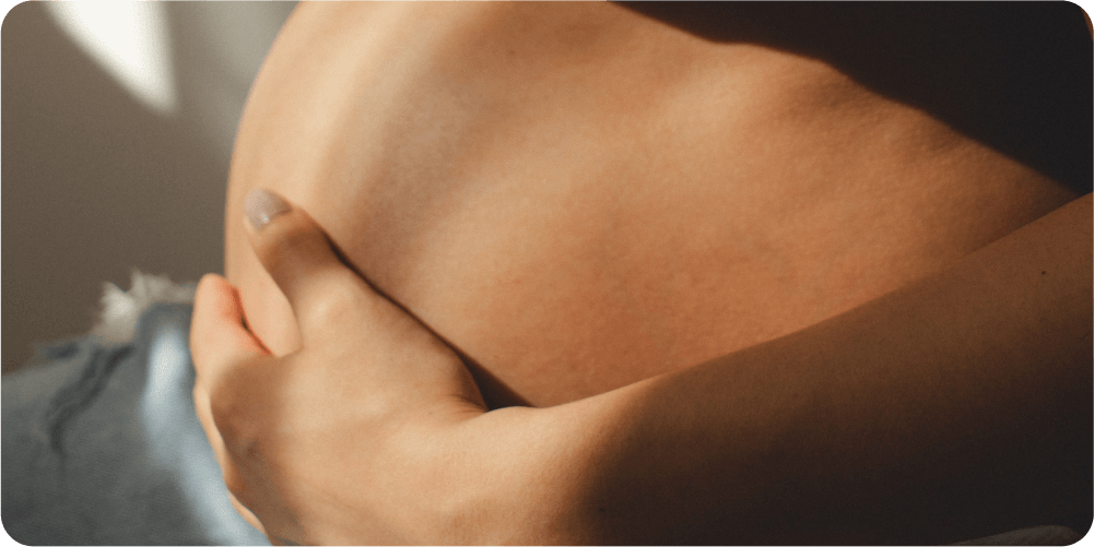 Крупный план беременного живота, нежно поддерживаемого рукой, залитый мягким естественным светом. Изображение передает тепло, нежность и материнскую заботу.
