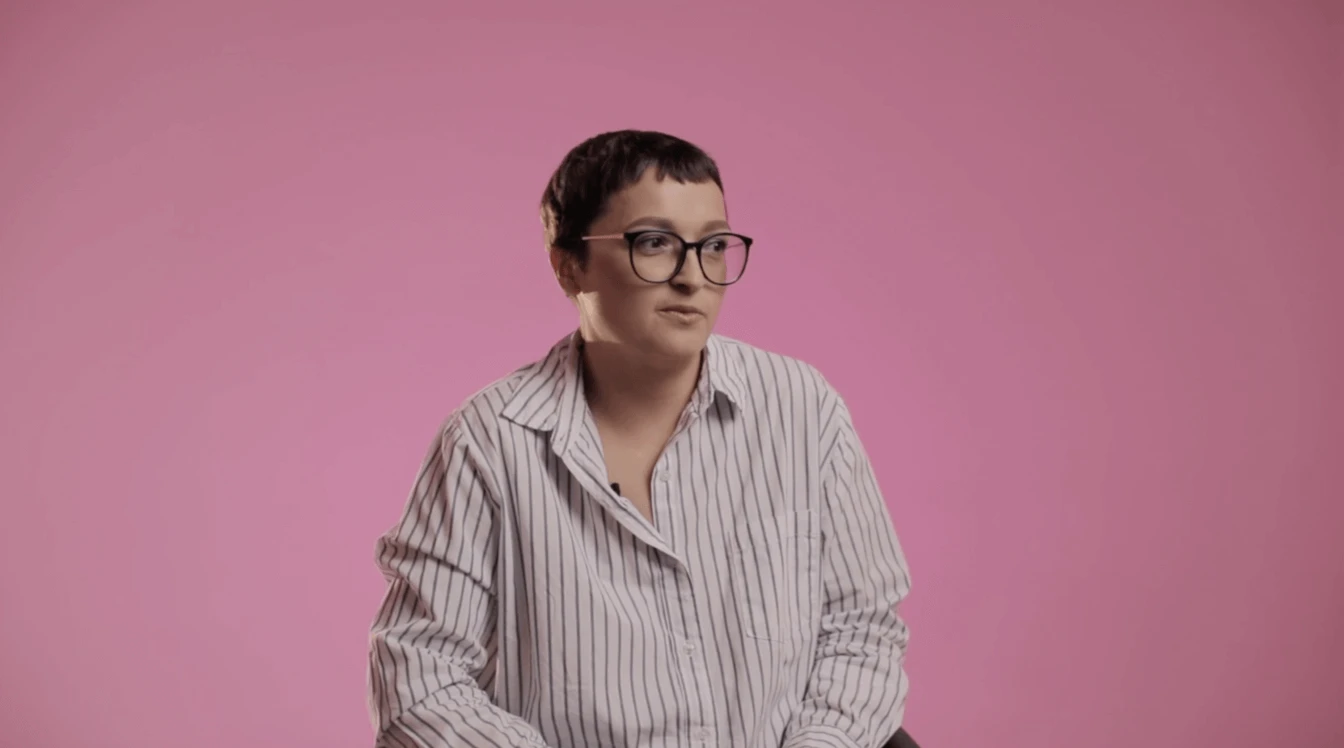 На розовом фоне сидит человек с короткой стрижкой, в очках и полосатой рубашке. Он выглядит задумчивым и расслабленным.