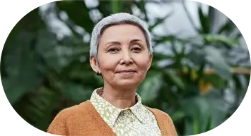 Пожилая женщина с короткими седыми волосами мягко улыбается на фоне пышных зеленых растений. На ней коричневый кардиган и узорчатая блузка, излучающие спокойствие и тепло.