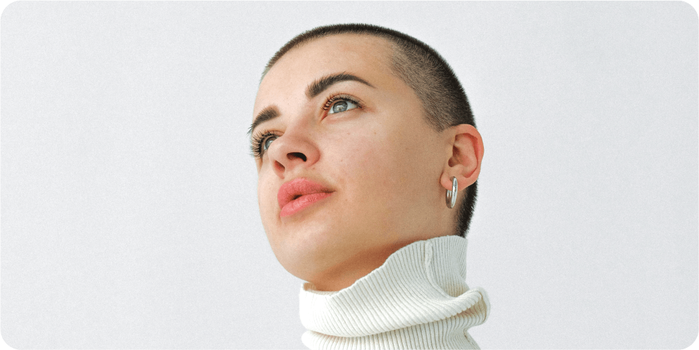 Человек с бритой головой и серьгами-кольцами смотрит вверх с безмятежным выражением лица. На нём белая водолазка на однотонном фоне.