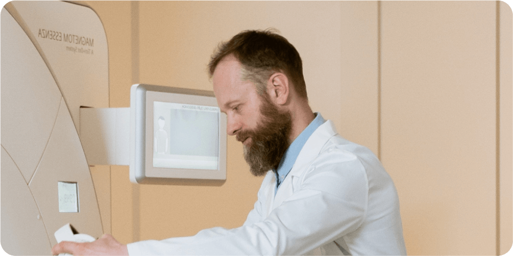 Врач в белом халате настраивает медицинское оборудование рядом с пациентом в клинике. Нейтральные тона помещения создают спокойную и профессиональную атмосферу.