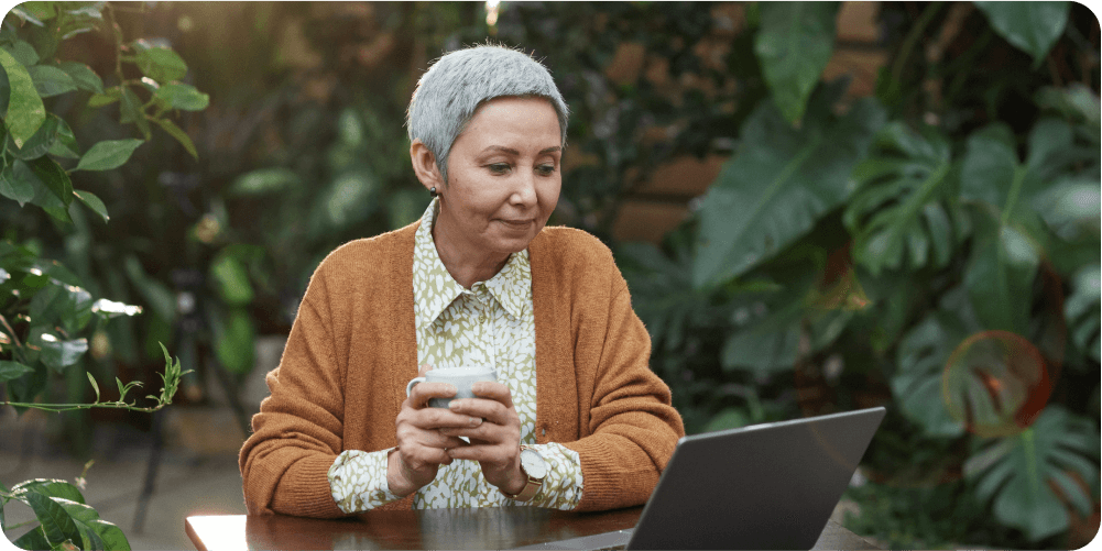 Пожилая женщина с короткими седыми волосами, в оранжевом кардигане, сидит за деревянным столом с чашкой кофе в руках и смотрит в ноутбук. Её окружают пышные зелёные растения.