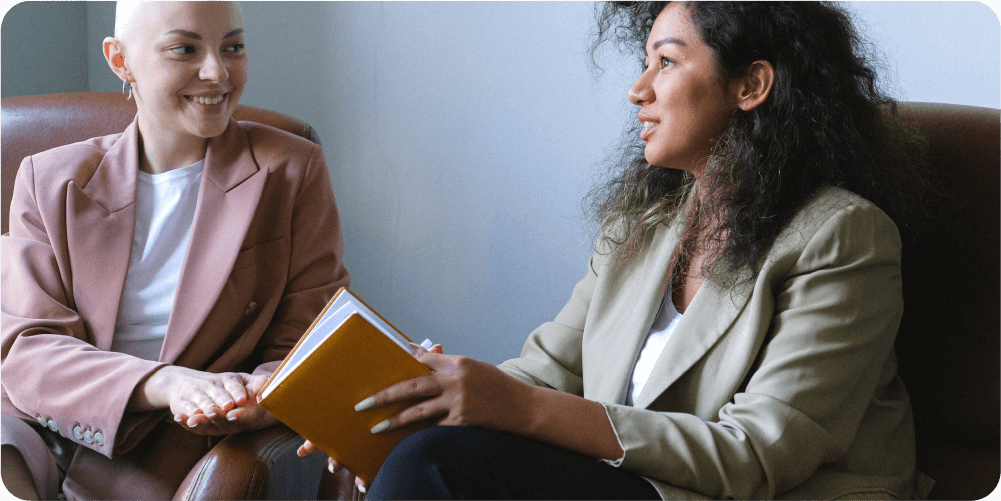 Двое сидят в креслах и ведут беседу. Один держит жёлтый блокнот. Оба выглядят расслабленными и улыбаются. Тёплая, деловая обстановка.