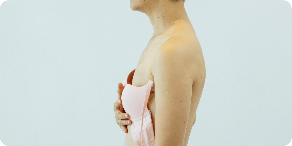 Человек с короткими волосами, стоящий в профиль, прижимает розовый бюстгальтер к обнаженной груди на простом светлом фоне, передавая уязвимость и самоанализ.