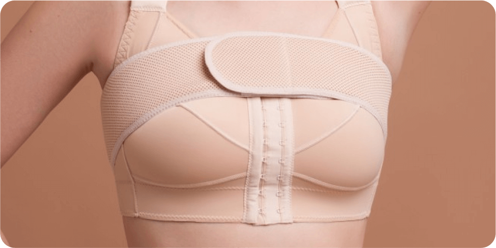 На бежевом фоне изображена женщина в бежевом послеоперационном компрессионном бюстгальтере. Бюстгальтер оснащен поддерживающей передней бретелькой и застежкой на крючки, что обеспечивает комфорт и поддержку.