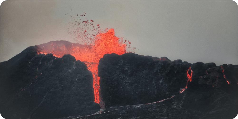 Из вулкана извергается лава, ярко-красные расплавленные породы вырываются на фоне тёмного, дымного неба. Сцена передает невероятный жар и природную мощь.